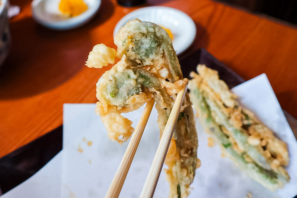 Chopsticks lift crispy vegetable tempura over a paper-lined plate on a wooden table, with dipping dishes blurred in back.