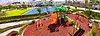 Equipamiento para Desarrollo Urbano Jogare. Vista panorámica de una zona recreativa infantil al aire libre con juegos coloridos (resbaladillas, columpios y estructuras trepadoras) sobre un suelo de caucho rojo. Al fondo se observa una cancha deportiva azul, áreas verdes y edificios residenciales modernos.