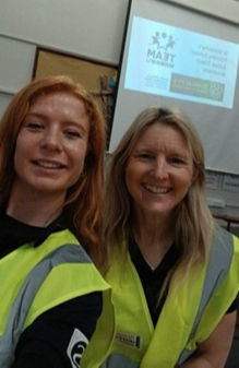 Two women who are Modeshift staff are smiling, wearing yellow safety vests in a classroom; a presentation slide is visible in the background.