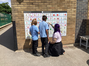 Two children standing outside with teacher, all facing a communication board