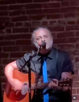 King seated, playing guitar and singing in front of a brick wall. Black shirt, blue tie. 
