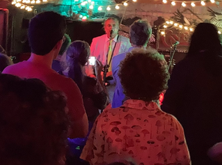 The King Teen in a seersucker suit, singing at the mic and playing guitar, seen from the back of the crowd. 
