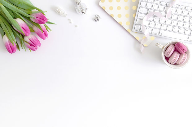 White office desktop with purple tulips, pearls, silver bird, gold and white polka dot mouse pad, white keyboard, white ribbon, and white coffee mug full of pink macaroons