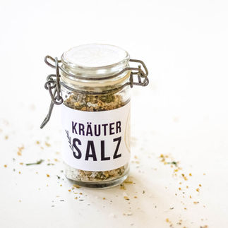 Kleiner Glasbehälter mit selbstgemachtem Zitronen Kräutersalz mit einer weissen Etikette wo drauf steht: Kräutersalz. Auf dem Deckel ist eine Etikette mit einer Illustration von einer gelben Zitrone