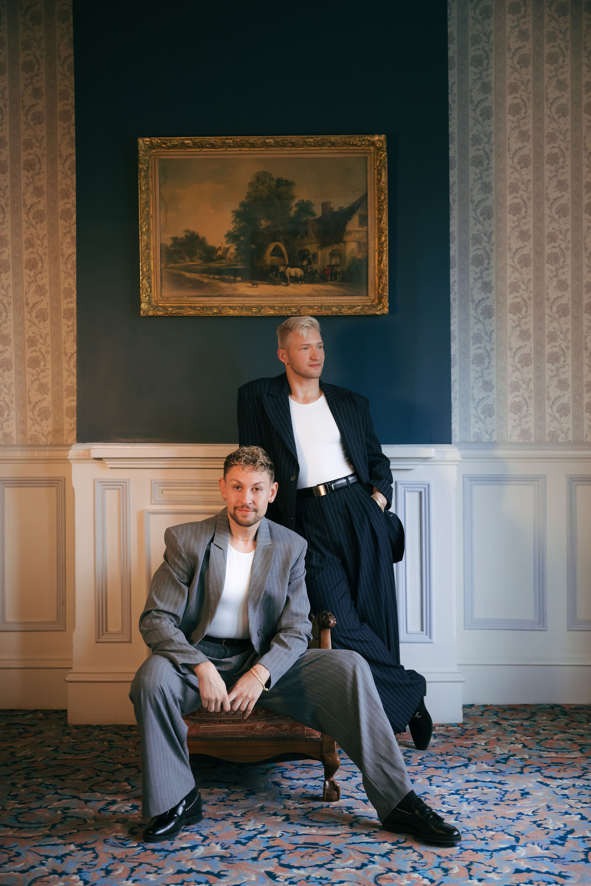 two men posing in front of a painting, one seated and the other leaning behind him