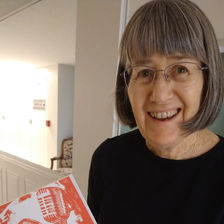 An older woman with brown hair and glasses holds an orange book
