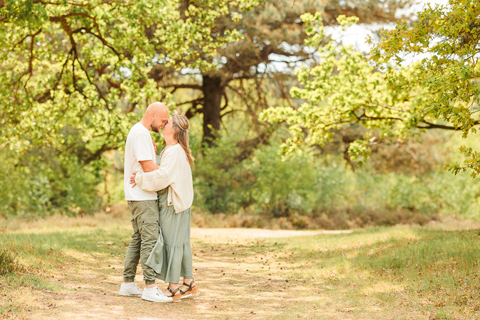 Verloofd stel tijdens een loveshoot in het bos, voorhoofden tegen elkaar in hard zonlicht op een groen pad.