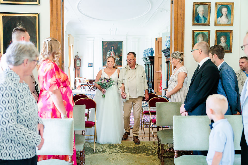 Bruid loopt gangpad op met haar vader in klassieke trouwzaal, terwijl gasten toekijken tijdens ceremonieel moment.