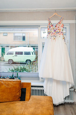 Kleurrijke trouwjurk van de bruid voor het raam, met de VW T3 buiten geparkeerd