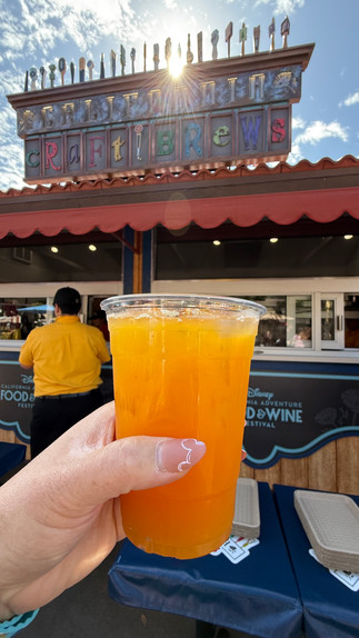 A close-up of a vibrant orange cocktail from the 2025 Disney California Adventure Food & Wine Festival, held in front of a beautifully decorated festival booth. The drink appears refreshing and fruity, served in a clear plastic cup with ice, capturing the golden glow of the sun. A perfect festival sip for those looking to enjoy a chilled, flavorful beverage while strolling through Disney California Adventure!