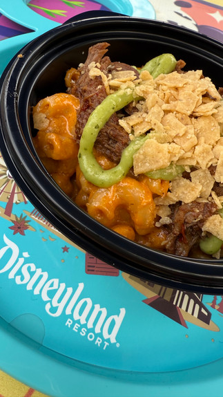 A close-up of the Birria Mac & Cheese from the 2025 Disney California Adventure Food & Wine Festival. This indulgent dish features creamy, rich mac & cheese topped with tender, slow-cooked birria beef, crispy tortilla strips, and a hint of spice for the perfect comfort food twist. Served in a black festival bowl on a vibrant Disneyland Resort plate, this flavorful fusion is a must-try for mac & cheese lovers and birria enthusiasts alike!