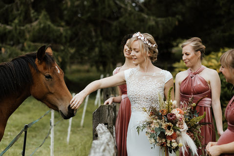 A bride dressed in a boho style dress with a jeweled headband in her hair is accompanied by her bridesmaids in burnt ochre colour, halter neck style dresses. They are looking at and patting a brown horse. 