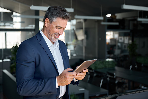 Happy mid aged business man ceo wearing blue suit standing in office using digital tablet