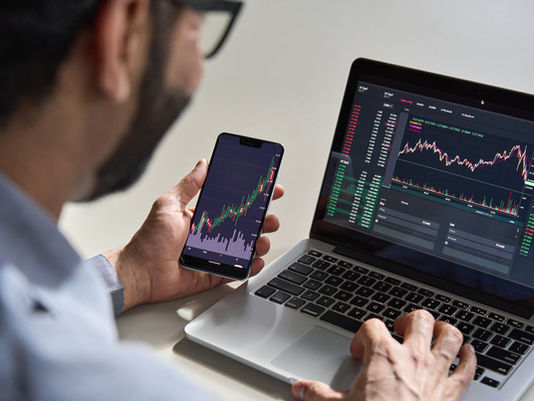 Person analyzing crypto charts on a smartphone and laptop. Screens display colorful graphs and data