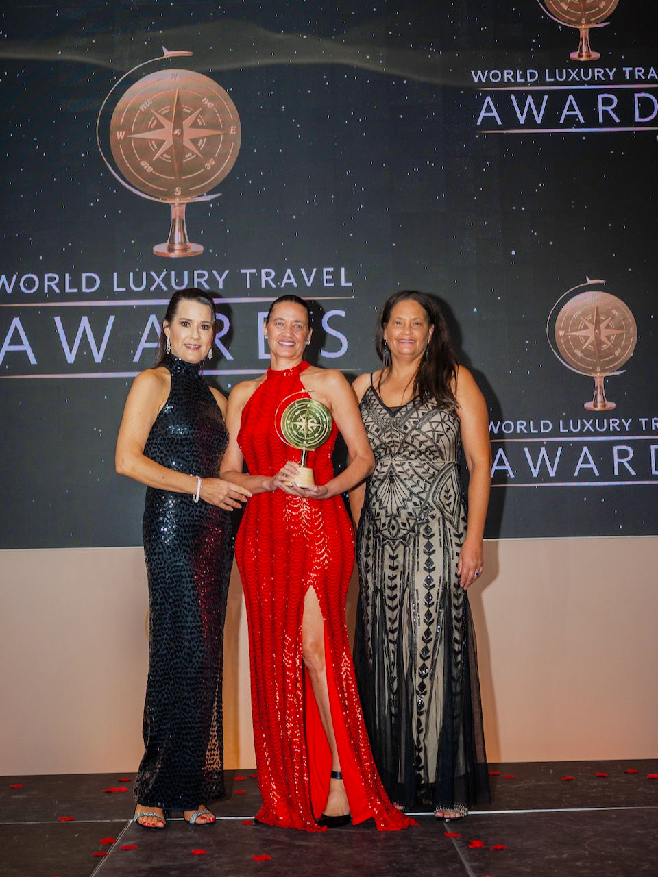 Villa Team in elegant dresses on stage, receiving an award. from the "World Luxury Travel Awards" text. Celebratory mood.