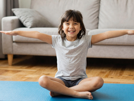 Bambina sorridente seduta a gambe incrociate su un tappetino blu, con le braccia aperte durante un esercizio di yoga in casa.