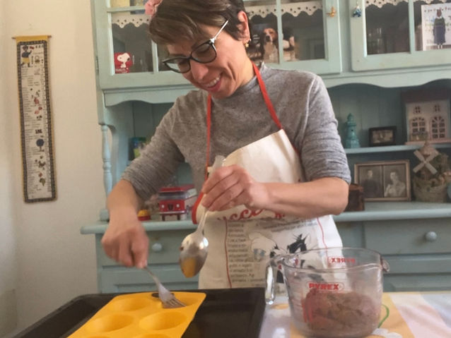 Donna sorridente con grembiule mentre prepara dei dolci in cucina, versando l’impasto in uno stampo di silicone giallo.