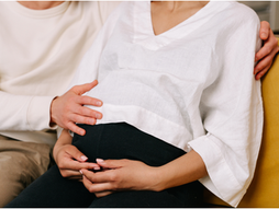 A couple sitting together.  The woman is pregnant.  Her partner has one arm around her and is feeling her belly with their other hand..