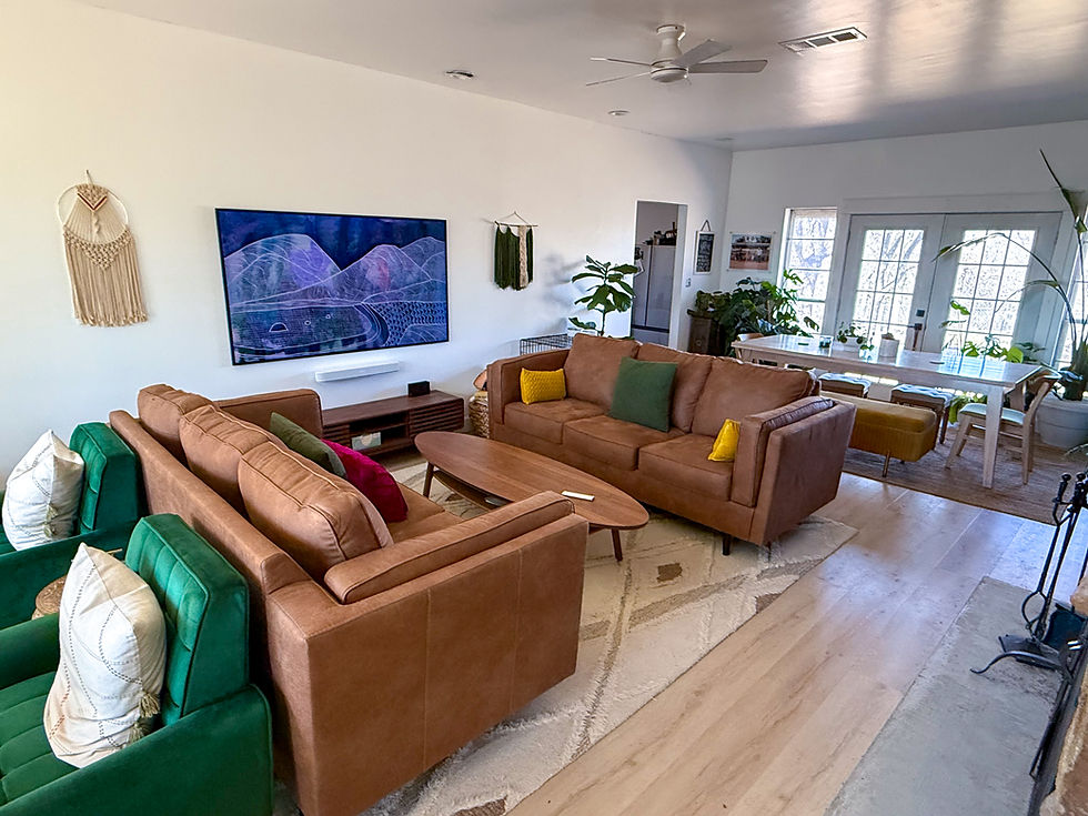 Cozy living room with brown sofas, green chairs, and wall art. Plants and a dining table in the background. Light, airy atmosphere.