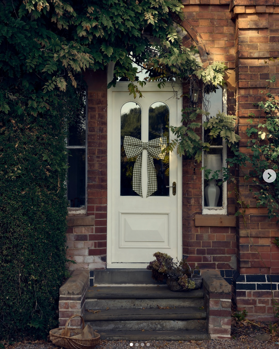 White door with large checkered bow, set in a brick wall covered with greenery. Basket of items on the steps, rustic and inviting mood.