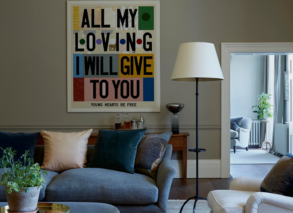Cosy living room with a grey sofa, colorful pillows, a floor lamp, and a wall art with text "ALL MY LOVING I WILL GIVE TO YOU." Bright ambiance.