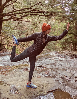 A woman with long brown, plaited hair, is in a Dancer's Pose in the middle of a wide stream that runs through a forest. She wears dark green leggings, and black quited coat and an orange beanie hat.