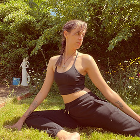 A female yogi is in Pigeon Pose in a sunlit garden lined with spring flowers. She wears black joggers and a cropped black top. She has brown hair, accented with purple, that's tied into a messy plait.