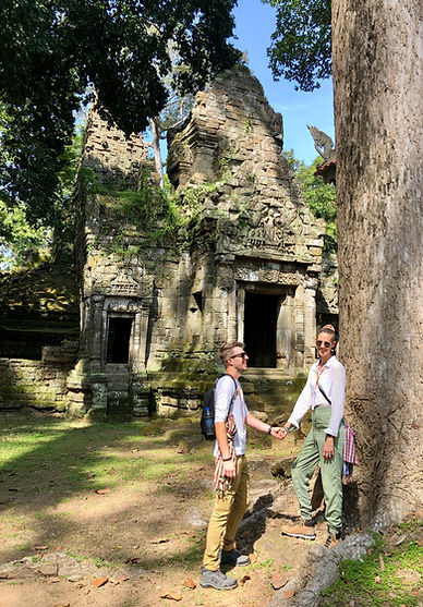 A young couple pose hand in hand in front of the ruins of Preah Palilay, Angkor, Cambodia. They both wear khaki trousers and and white tops, with checked scarfs tied at their waists. The ruins are lined with moss and tree roots.