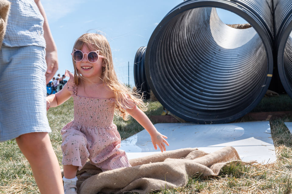 Young girl runs toward large tubes; smiles, wearing sunglasses. Thompson Adventure Farm setting.