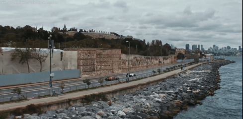 Two Formula 1 cars drive along the coastal road beside the Sarayburnu walls in Istanbul, with the rocky shoreline and choppy water on one side and the historic palace walls rising above the road on the other. The wide view connects modern motorsport with the layered waterfront landscape of Sarayburnu.
