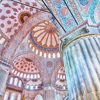 Upward view inside the Blue Mosque showing layered domes, painted arches, blue tilework, and a massive stone column rising through the prayer hall. The composition emphasizes the mosque’s ornate ceiling patterns and monumental interior scale.