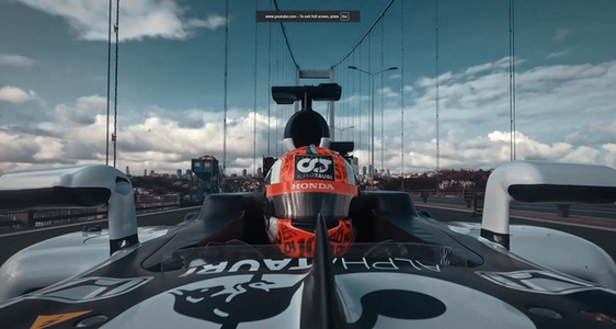 A Formula 1 car drives across the Bosphorus Bridge in Istanbul, seen from above with suspension cables rising vertically and the water stretching below on both sides. Hills packed with buildings fill the background