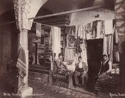 Historic photo of a fabric shop in the Grand Bazaar with merchants sitting in front of hanging textiles.