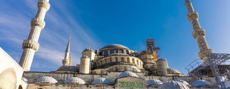 Front view of the Blue Mosque with layered gray domes and six slender minarets rising above the courtyard arcade under a bright blue sky with scattered clouds. The low angle emphasizes the symmetry and monumental scale of the mosque’s exterior.