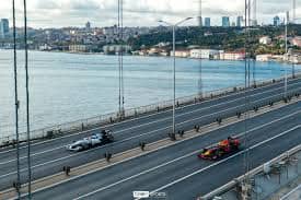 Two Formula 1 cars drive across a bridge in Istanbul with the Bosphorus and city buildings visible in the background. The image highlights the contrast between high speed motorsport and Istanbul’s urban waterfront setting.