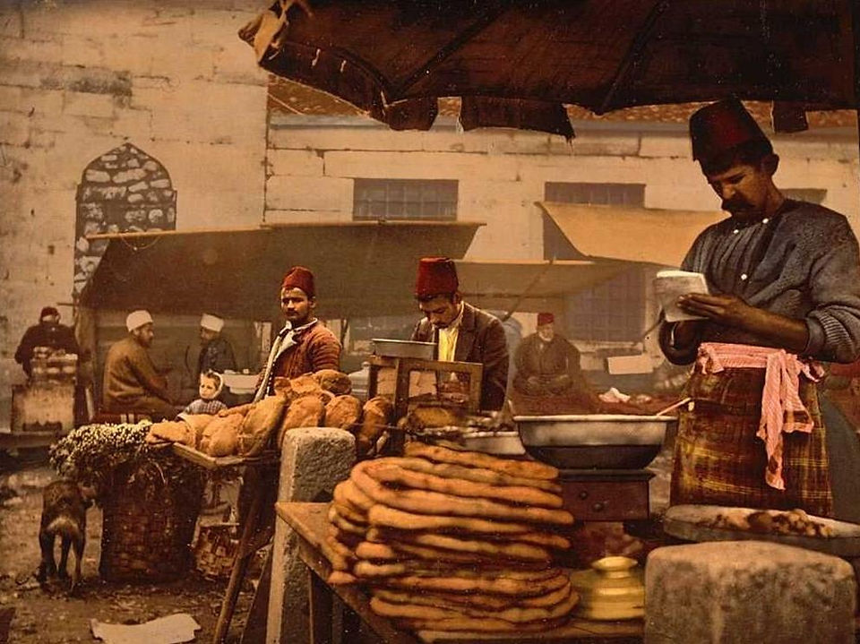 A historical depiction of an Ottoman-era open-air market with men in traditional attire selling bread and street food.