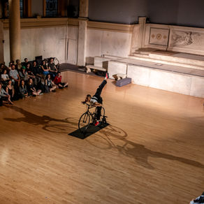 Banner image featuring Katherine on the bike at Judson Memorial Church. She balances on one pedal while the other leg extends in a penché behind her. Audience members watch seated at either side. Photo by Rachel Keane.