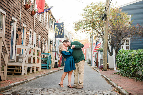 A man dressed in a green sweater and light trousers romantically dips a woman wearing a teal dress on a charming street in Annapolis. Surrounded by historic buildings, they share a passionate kiss, creating a timeless and enchanting moment.