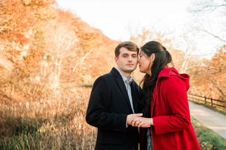  In the midst of a vibrant autumn forest scene at Great Falls State Park in Virginia, a man and woman, dressed in sophisticated black attire, share a tender moment. Against the backdrop of trees adorned with orange leaves, the woman's bright red jacket adds a pop of color to the serene landscape. Their closeness is evident in the subtle intertwining of their fingers and the softness of their gaze, reflecting the depth of their connection amidst the natural beauty surrounding them. This intimate portrait captures the warmth of their love and the anticipation of their journey together amidst the enchanting fall foliage.