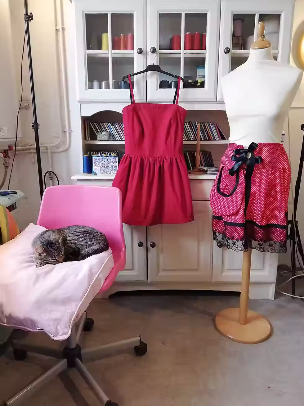 atelier doux c'est une photo de créations personnalisées pour une cliente dans les tons rouges/ rose avec le buffet plein de bobines de fils multicolore en arrière plan, la machine industrielle sur le coté en au centre la chaise rose où dort tranquillement le chat