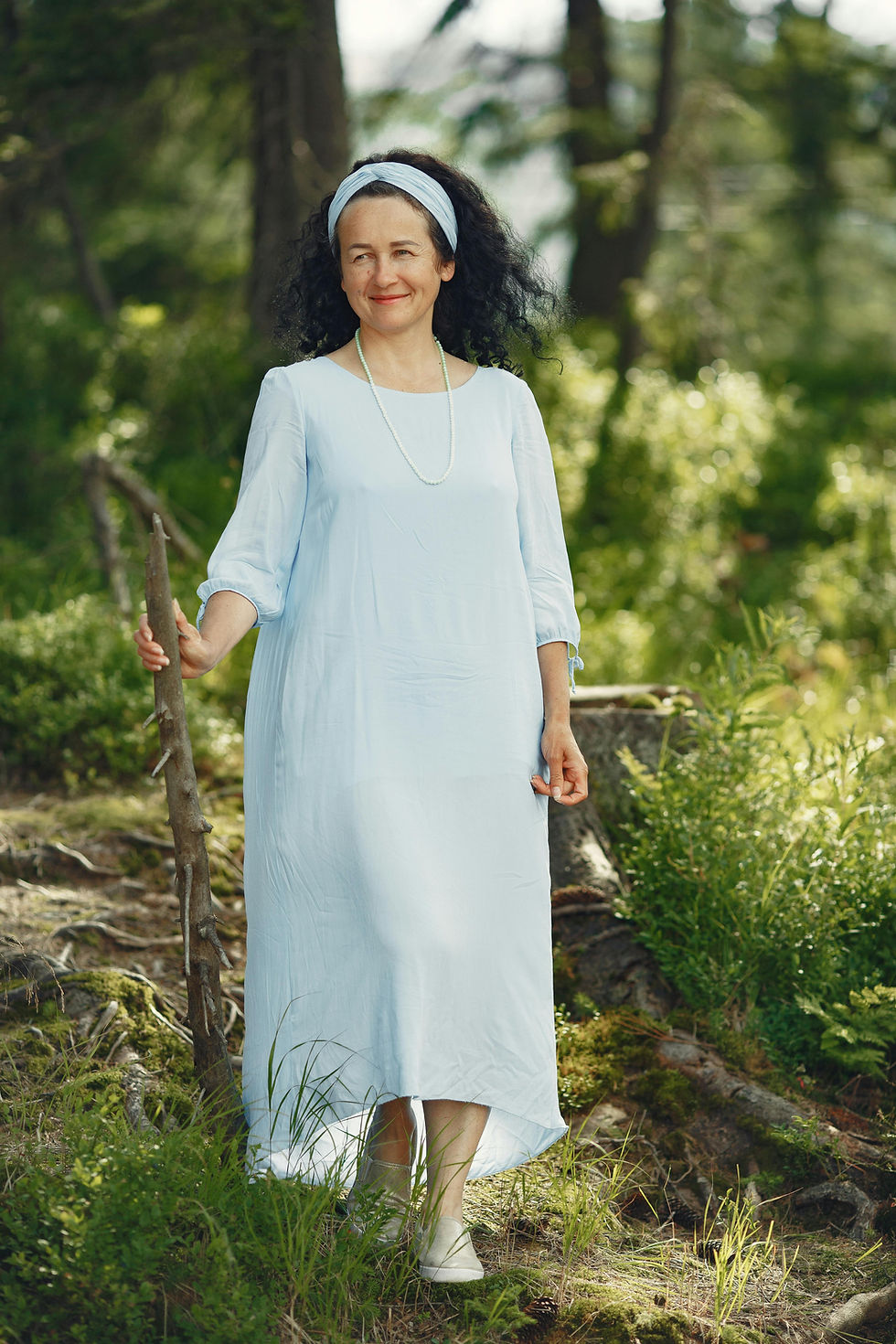 une femme de cinquante ans qui porte une robe ample et confortable en coton tout en restant féminine, elle porte un bandeau dans les cheveux elle a de beaux cheveux longs et bouclés, elle marche dans la nature en tenant un bâton elle sourie, c'est l'image même d'une druidesse du 21 ème siècle rayonnante