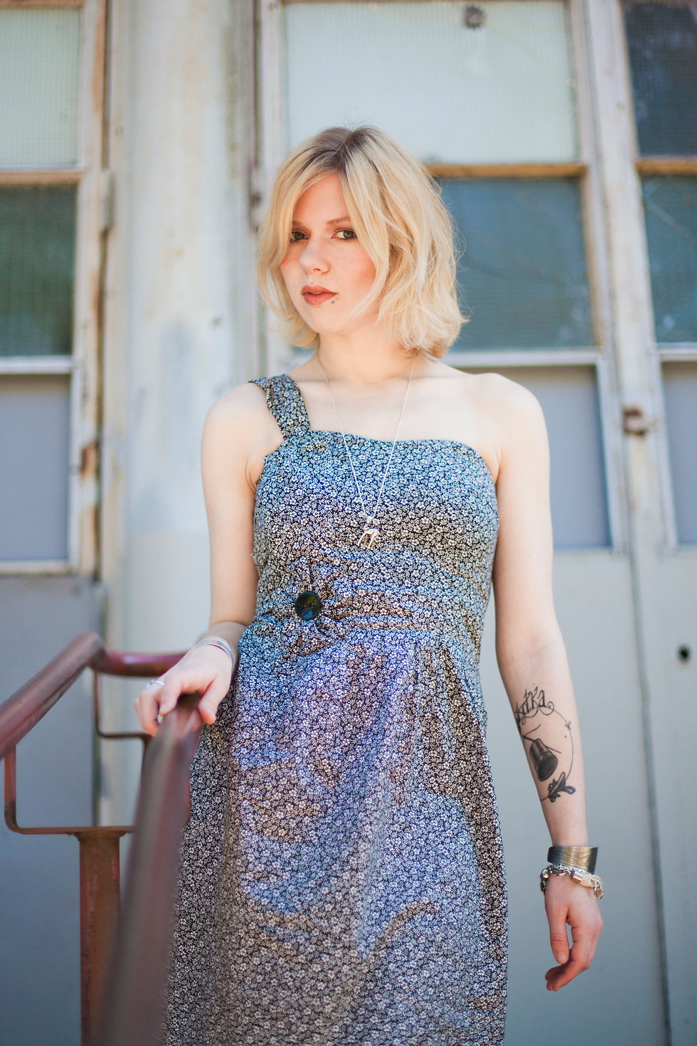 une jolie femme blonde avec un carré pixie et des lunettes de soleil ray ban qui ressemble à scarlett johanson qui porte une robe one shoulder de créateur français en tissu popeline de coton liberty à fleurs