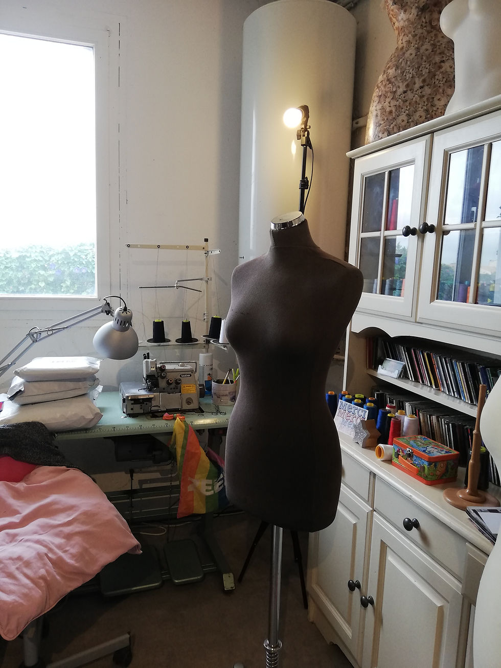 Photo de la machine à coudre industrielle, une surjeteuse sur laquelle repose des colis de tissu à déballer, devant il y a un mannequin de couture noir et sur le coté droit le buffet qui contient toute la mercerie de l'atelier