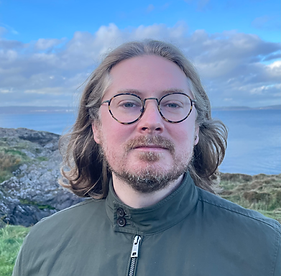 A picture of Daniel standing by the sea: long blode hair with flecks of grey, round glasses and a short beard. He is wearing a green jacket.