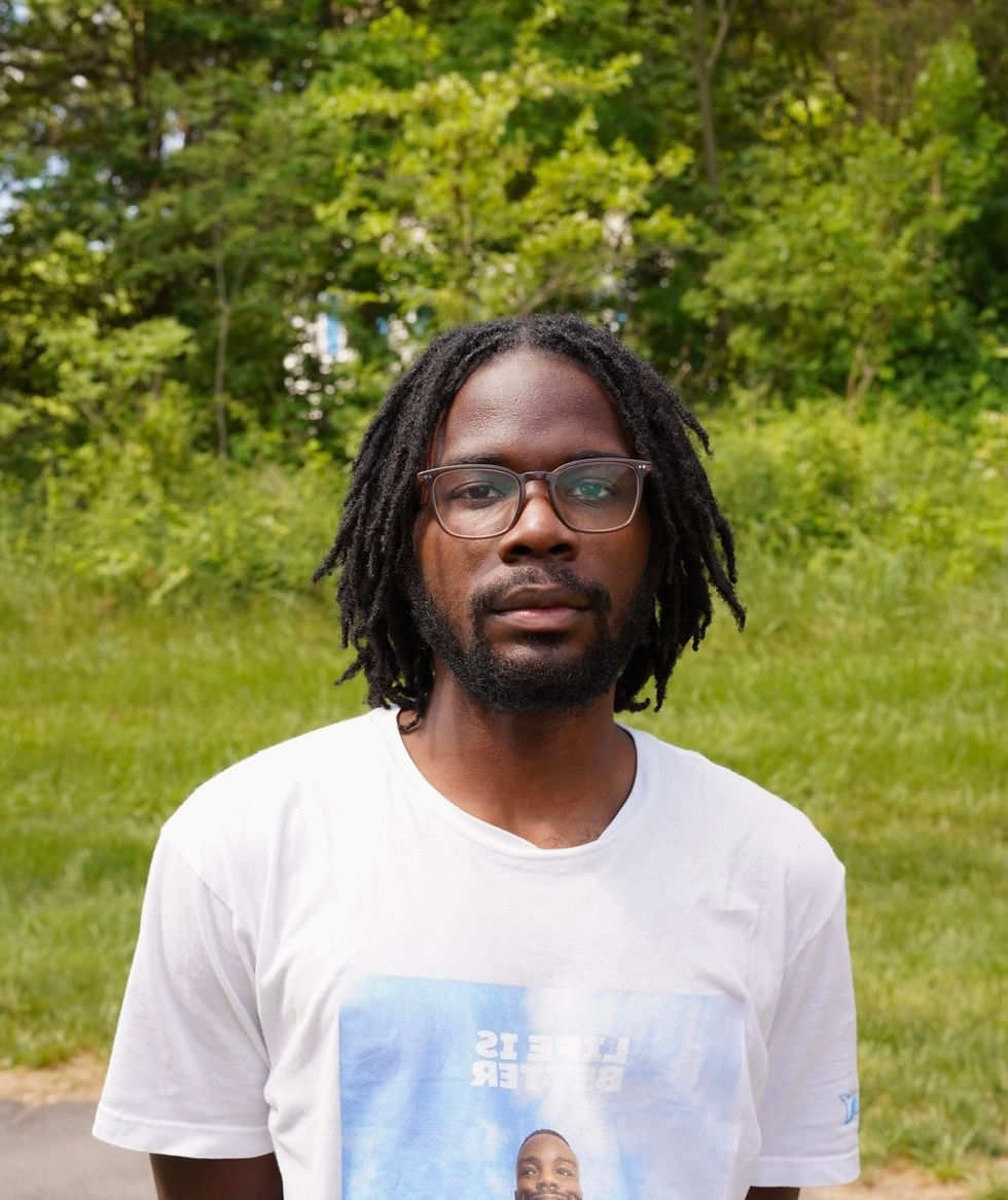 Fatimah Asghar in peach dress holds yellow rose, sitting amid vibrant flowers. Star earrings, henna tattoos, and ornate drapery create an artistic mood.