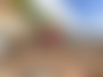 Two women stand together on a massive pile of shattered wood, metal, and debris—the remains of a landslide-destroyed building. One holds a worn American flag. Behind them, steep cliffs rise under a blue sky with scattered clouds