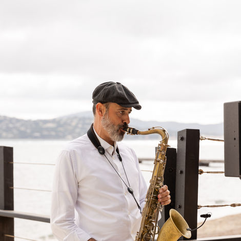 Saxophoniste jouant du saxophone, chemise blanche, chapeau, près de l'eau, performance musicale.
