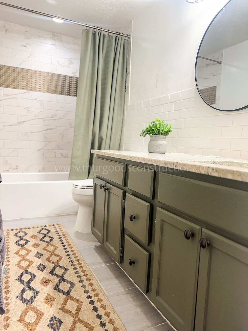 Bathroom with green cabinets, round mirror, subway tile, plant on counter. Beige patterned rug, green shower curtain, and white tub.