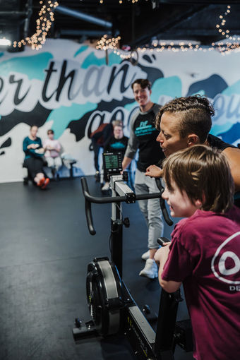 Woman and child smiling by exercise bike with 'er than' background.
