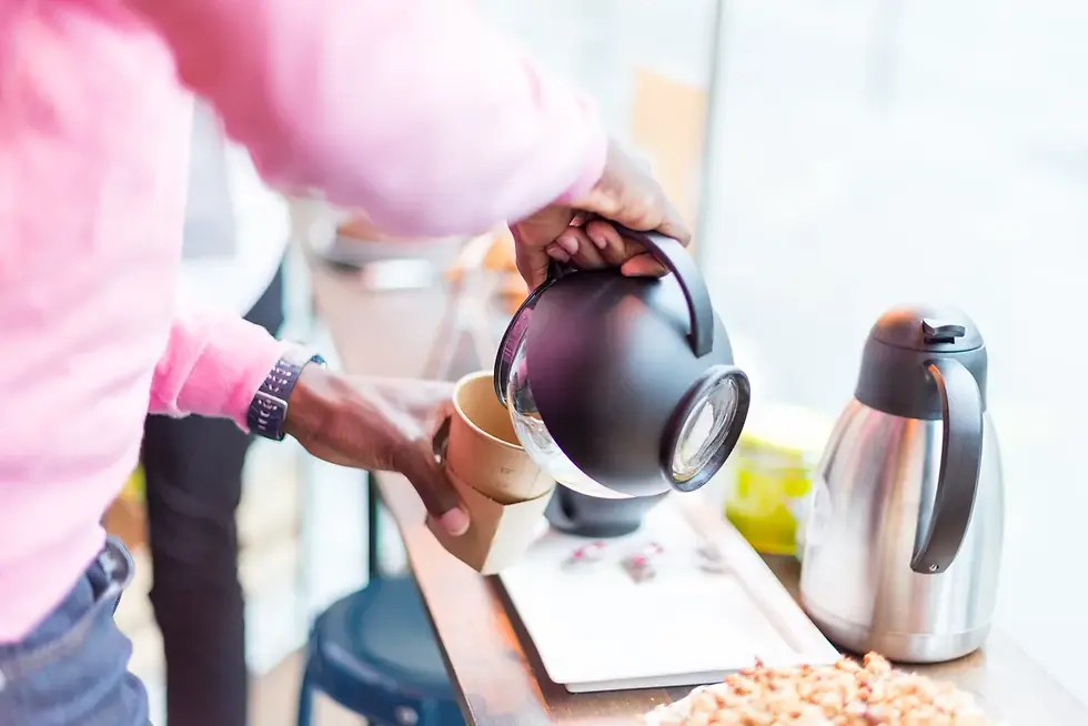 Photographe événementiel Aix-les-Bains - jeune homme en pull rose versant du café dans un gobelet, debout près d'un comptoir en bois avec cafetière argentée en arrière-plan.